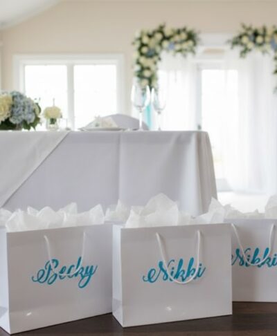Three personalised glossy white gift bags are arranged against a wedding-themed background dominated by light blue and white colors. The bags have white rope handles, and one bag, positioned centrally, is clearly labeled with the name "Nikki" in a flowing blue script font. The surrounding decor includes lush light blue and white floral arrangements and possibly some greenery, setting a sophisticated and romantic wedding party mood.