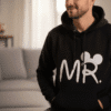 close up of a man sitting in a modern, brightly lit living room wearing a black hoodie. The hoodie features a large white graphic of the word "MR." where the "R" is styled with round mouse ears.