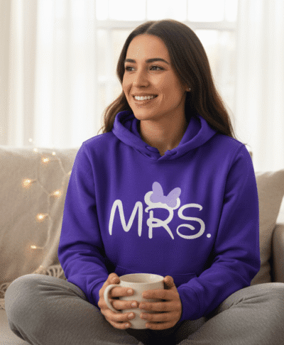 Lifestyle image of a woman sitting down and wearing a comfortable purple hoodie and trousers, clearly displaying the personalised white text "Mrs" on the front, ideal for bridal loungewear and personalised gifts.