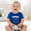 A joyful baby is sitting cross legged on a plush white rug, looking directly at the camera with a wide, happy smile. The baby is wearing a short-sleeved, bright blue bodysuit with white text and graphics that read: a mustache icon, "Little Groomsman Noah 30.06.23". The baby is holding a small, silver-wrapped gift box tied with a white ribbon in their hands. The background is softly blurred, showing a bright, airy space with white walls, soft lighting, and more silver-wrapped gifts and string lights, suggesting a festive or wedding related celebration.