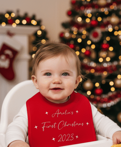 A close-up image of a baby wearing a red bib with a festive print. The background is softly blurred but has warm, cosy colors suggestive of a Christmas setting.