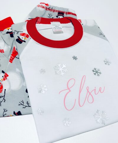 A set of children's Christmas pyjamas with a white raglan style top featuring a red collar and a printed snowflake design. The top is personalised with the name 