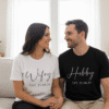 Lifestyle image of a newly married couple embracing in a bright, modern home setting, both wearing matching personalised t-shirts: the woman in a 'Wifey' tee and the man in a 'Hubby' tee, showcasing comfortable and cute couple apparel for newlyweds.